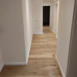 Jasny korytarz z nową podłogą z paneli drewnianych, białe ściany z listwami przypodłogowymi i oświetleniem punktowym w suficie.