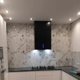 Nowoczesna kuchnia z białymi szafkami, czarnym okapem i marmurowymi płytkami na ścianie, oświetlona punktowymi lampami sufitowymi.