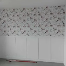 Ściana z dekoracyjnym panelem dolnym i tapetą w kwiaty. Czerwona poziomica leży na szarej podłodze.