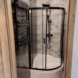 Marcin Rzeźnik - Nowoczesna kabina prysznicowa z czarnymi profilami i deszczownicą w wykończonej łazience. Na ścianach płytki w odcieniach szarości i beżu. Widoczny grzejnik drabinkowy.