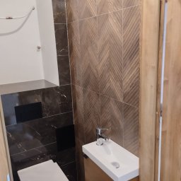 Marcin Rzeźnik - Nowoczesna toaleta z brązowymi płytkami w jodełkę i marmurowymi akcentami. Biała umywalka z szafką z naturalnego drewna. Minimalistyczny design, czarne elementy armatury.