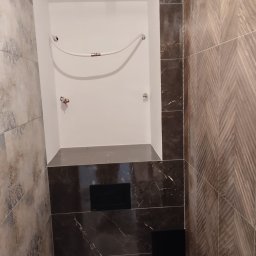 Marcin Rzeźnik - Narożne ujęcie wykończonej łazienki z zabudową WC, gdzie widoczne są płytki imitujące marmur i drewno oraz instalacja wodna w białej wnęce.
