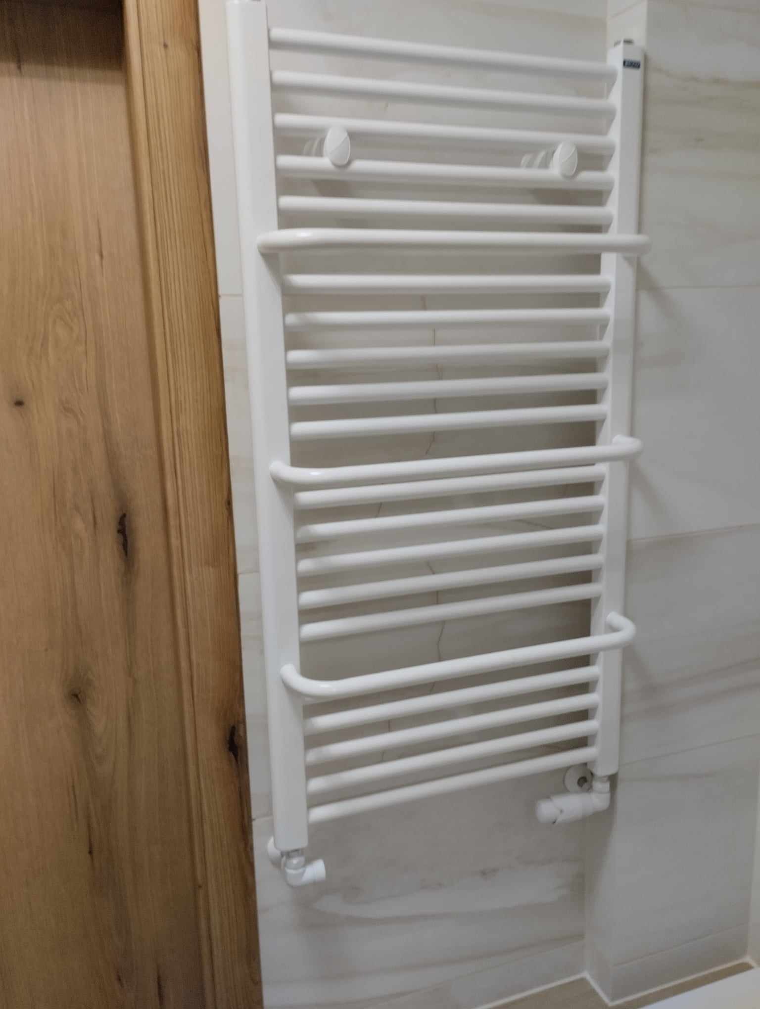 Biały, drabinkowy grzejnik łazienkowy zamontowany na ścianie z jasnych płytek, widoczny obok drewnianych drzwi.
