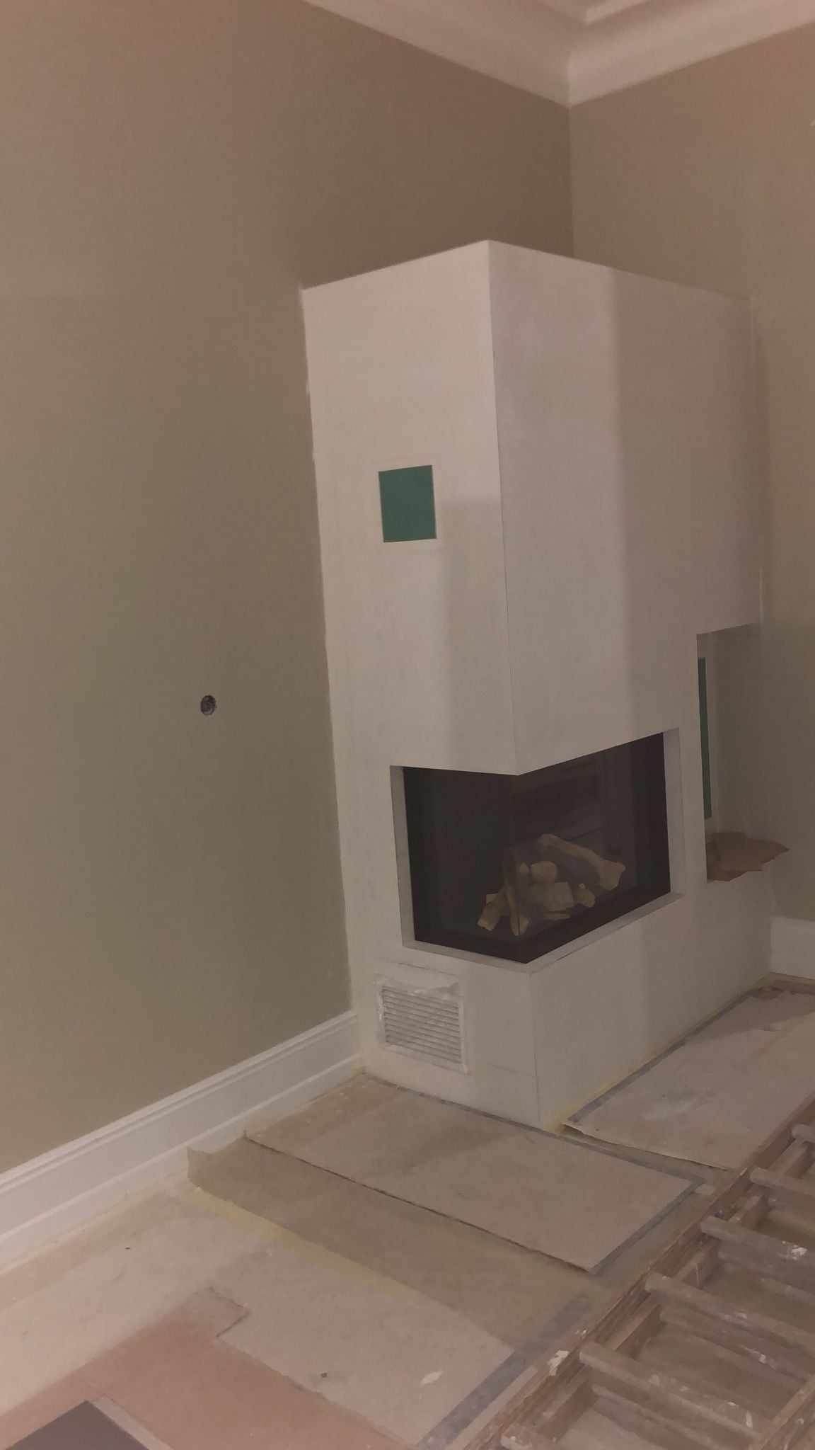 Kanciasty, biały kominek narożny z wkładem kominkowym w trakcie montażu, z widoczną zieloną naklejką zabezpieczającą, na podłodze płyty kartonowo-gipsowe i odsłonięta konstrukcja podłogi.