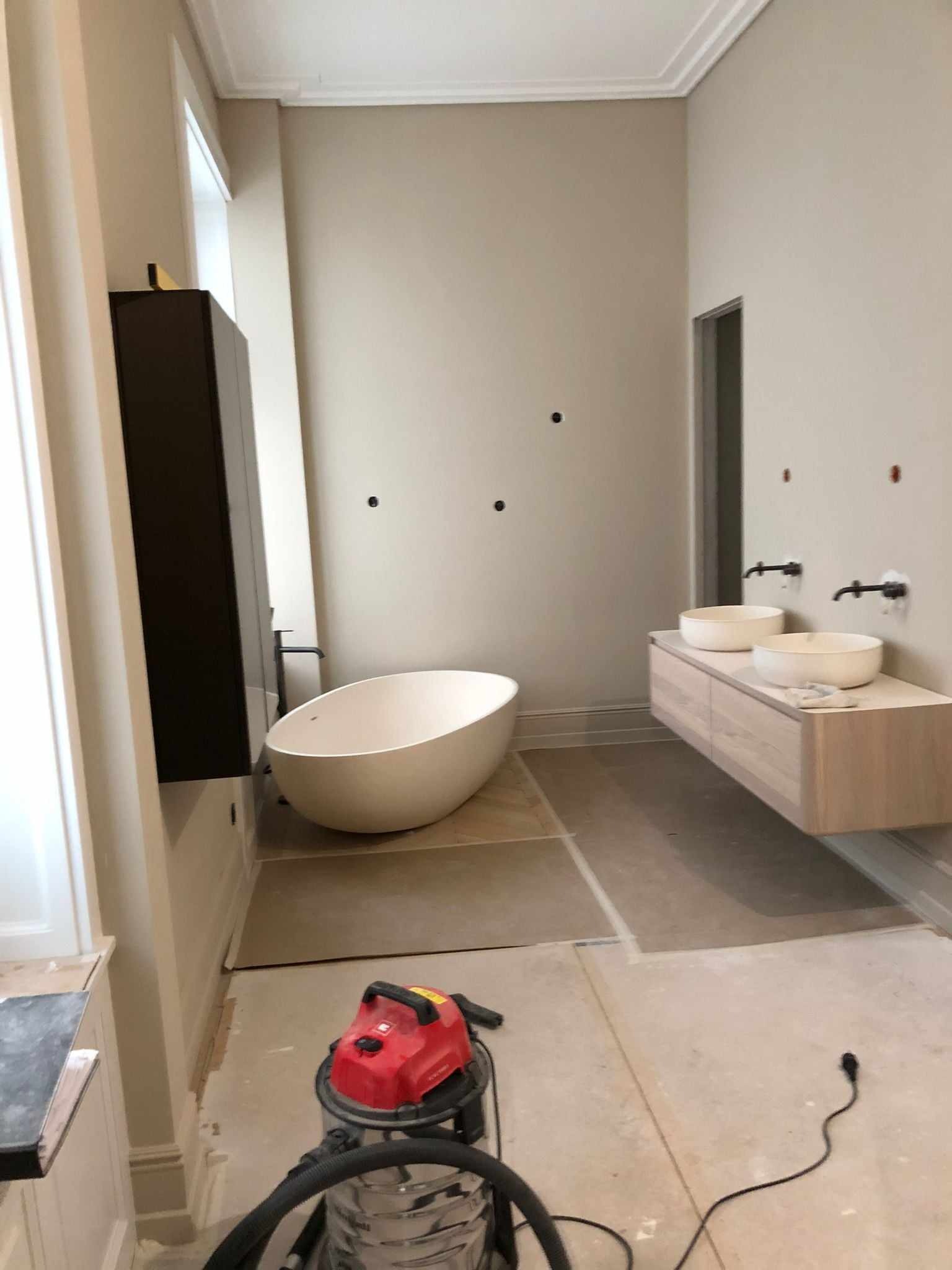 Łazienka w trakcie wykańczania: wolnostojąca wanna, podwieszana szafka z dwiema umywalkami nablatowymi, widoczny odkurzacz przemysłowy i przygotowane podłoże pod płytki.
