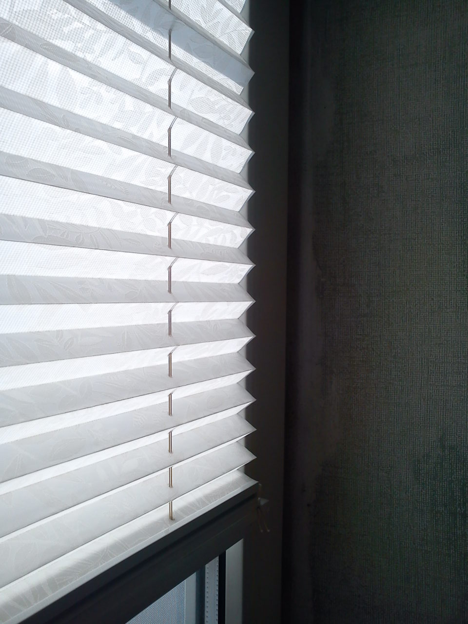 Białe plisy okienne z delikatnym wzorem, częściowo zasłonięte, widoczne przy oknie z ciemną ramą na tle ciemnej ściany z wyraźną teksturą.