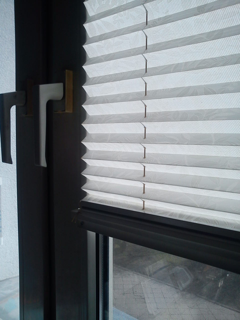 Zamknięte, białe plisy okienne z delikatnym wzorem liści na tle okna z moskitierą i czarną ramą.