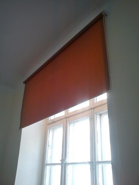 Pomarańczowa roleta wewnętrzna częściowo zasłania białe okno z firankami, widok z dołu.