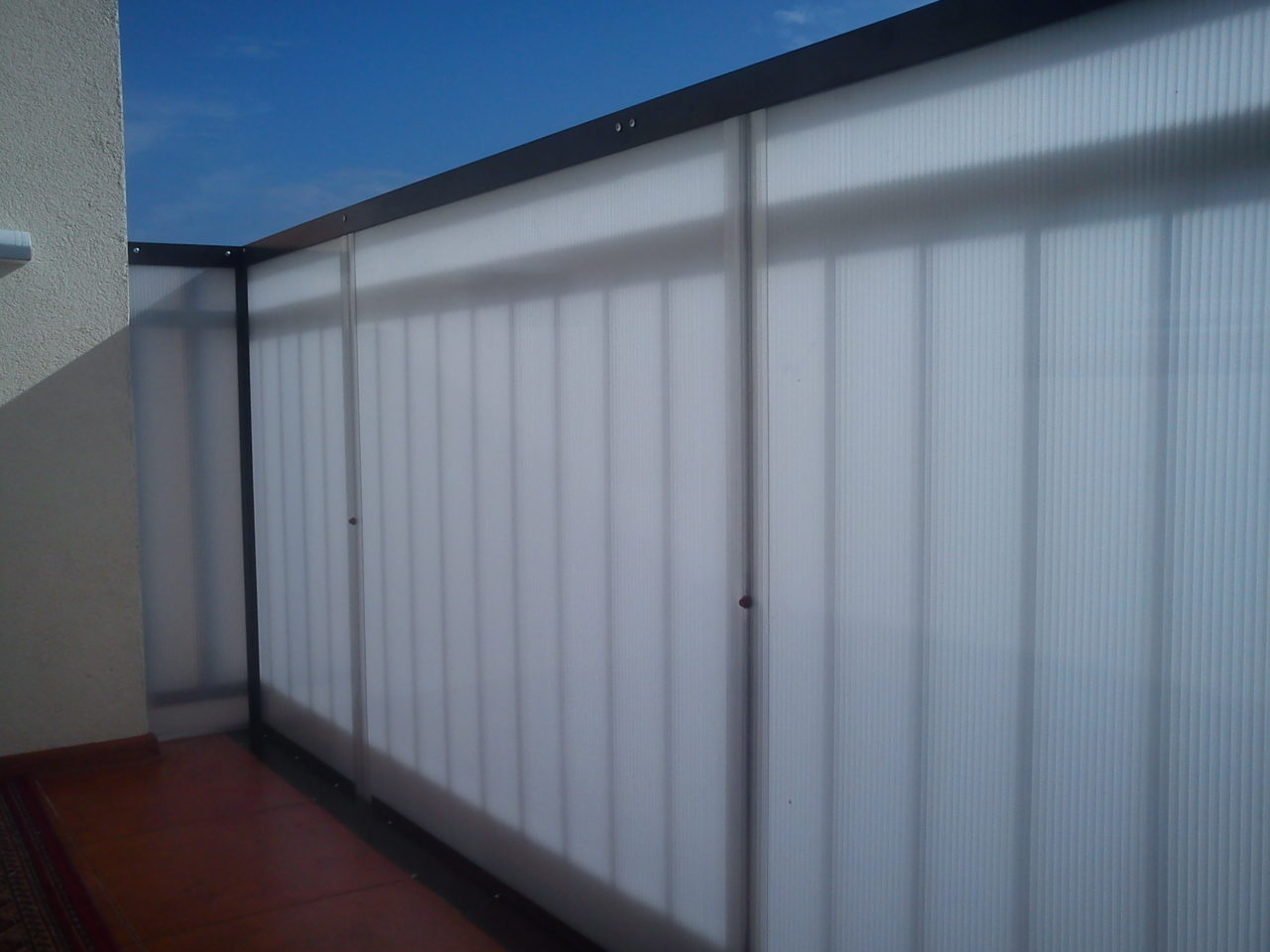 Zabudowa balkonu z półprzezroczystych paneli z ciemną ramą, widok z dołu na tle błękitnego nieba, z widoczną podłogą balkonu z płytek.