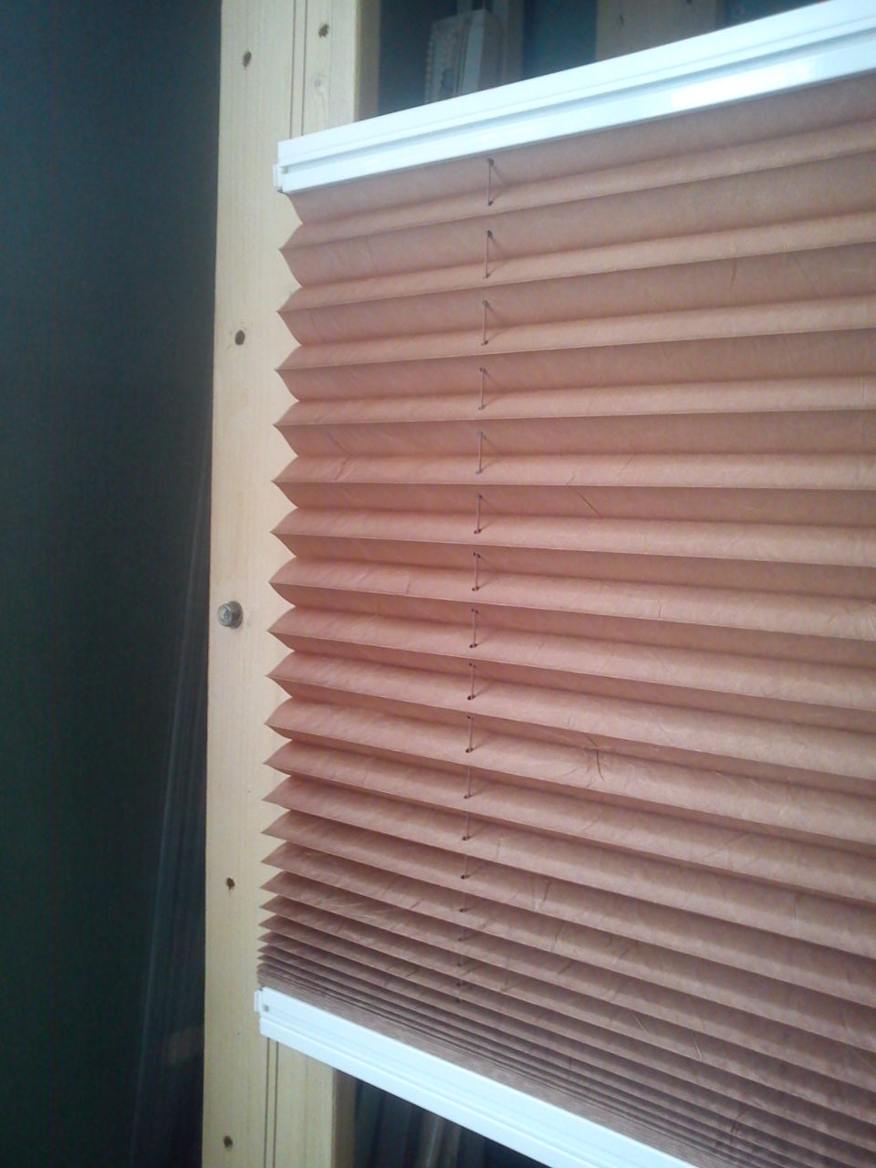 Beżowa, plisowana roleta okienna zamontowana na drewnianej ramie, widoczne sznurki regulujące wysokość.