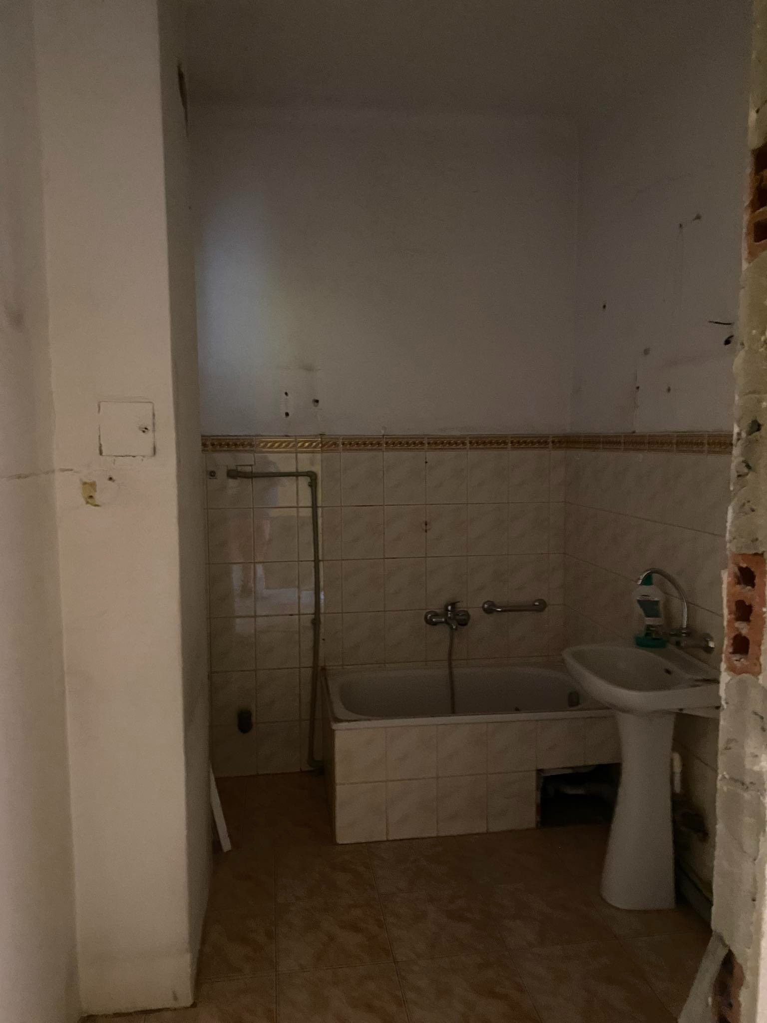 Łazienka w trakcie prac remontowych: widoczna wanna, umywalka na postumencie, stare płytki, ślady po skuciu tynku na ścianie i niezabezpieczone rury, sugerujące demontaż instalacji.