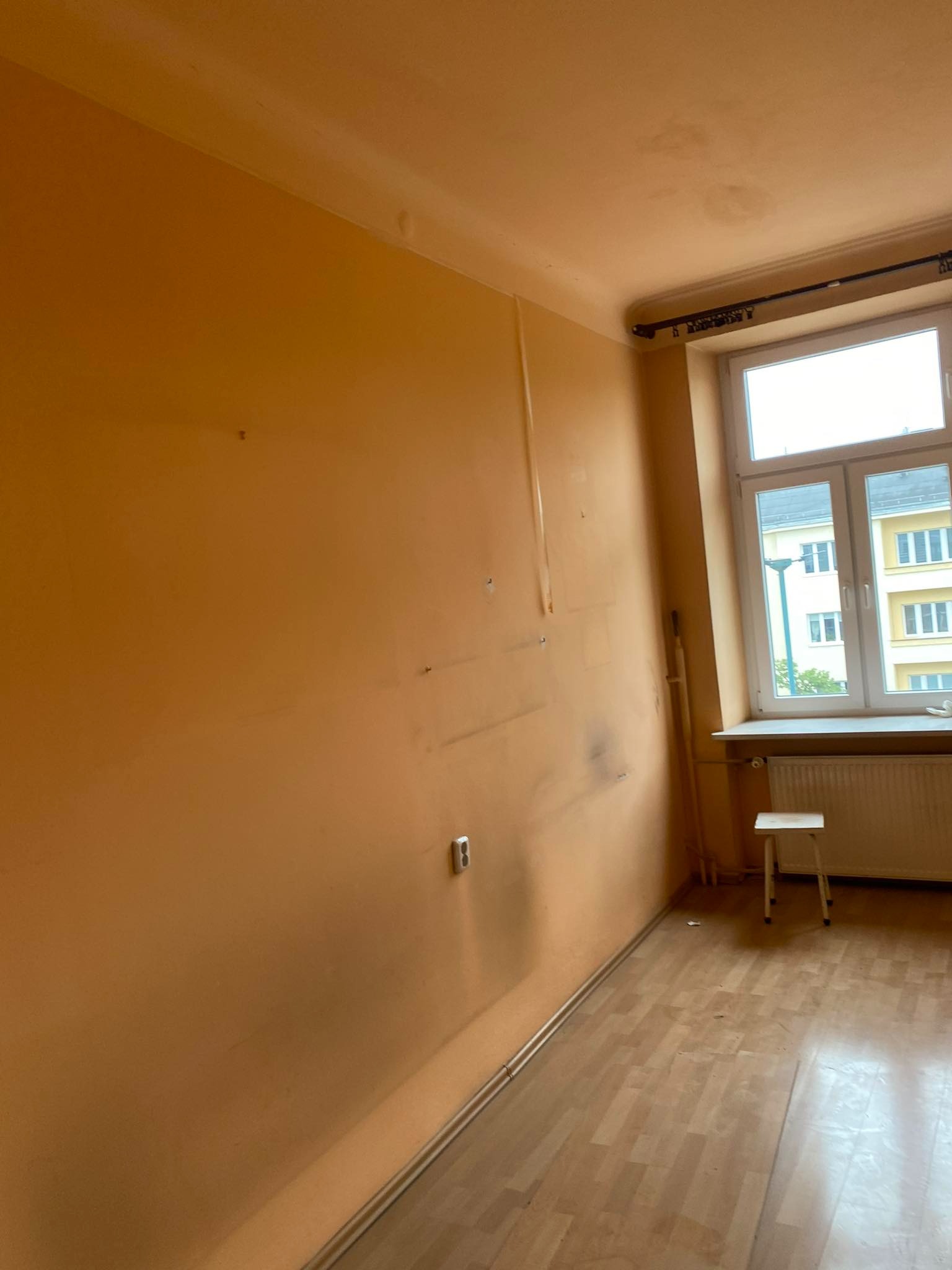 Pokój przed remontem: pomarańczowe ściany z widocznymi śladami po taśmie i otworach, drewniana podłoga, białe okno z widokiem na budynek, mały taboret przy kaloryferze.