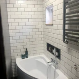 Biała narożna wanna obudowana białymi płytkami typu metro z ciemną fugą, grzejnik drabinkowy w kolorze grafitowym, wnęka w ścianie z półkami i kosmetykami, minimalistyczna armatura.