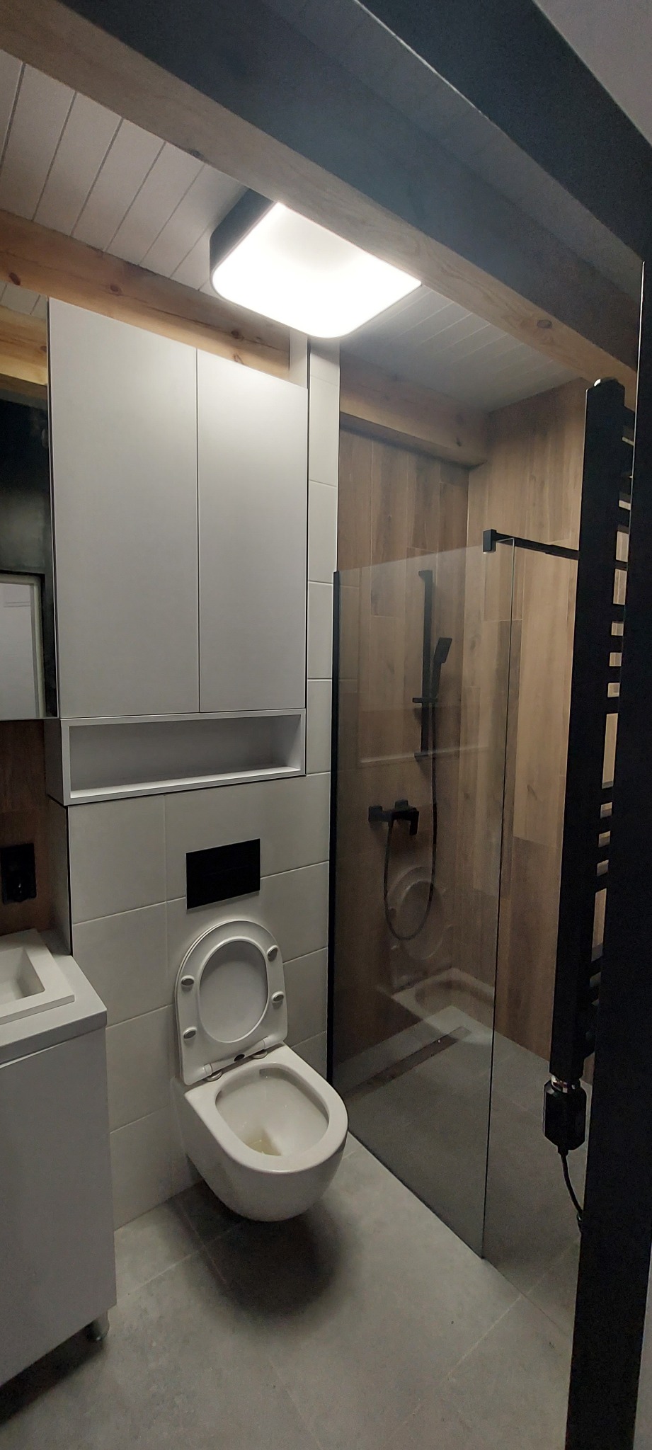 Nowoczesna łazienka z kabiną prysznicową typu walk-in, podwieszaną toaletą z zabudową i drewnianymi akcentami na suficie i ścianie prysznica.