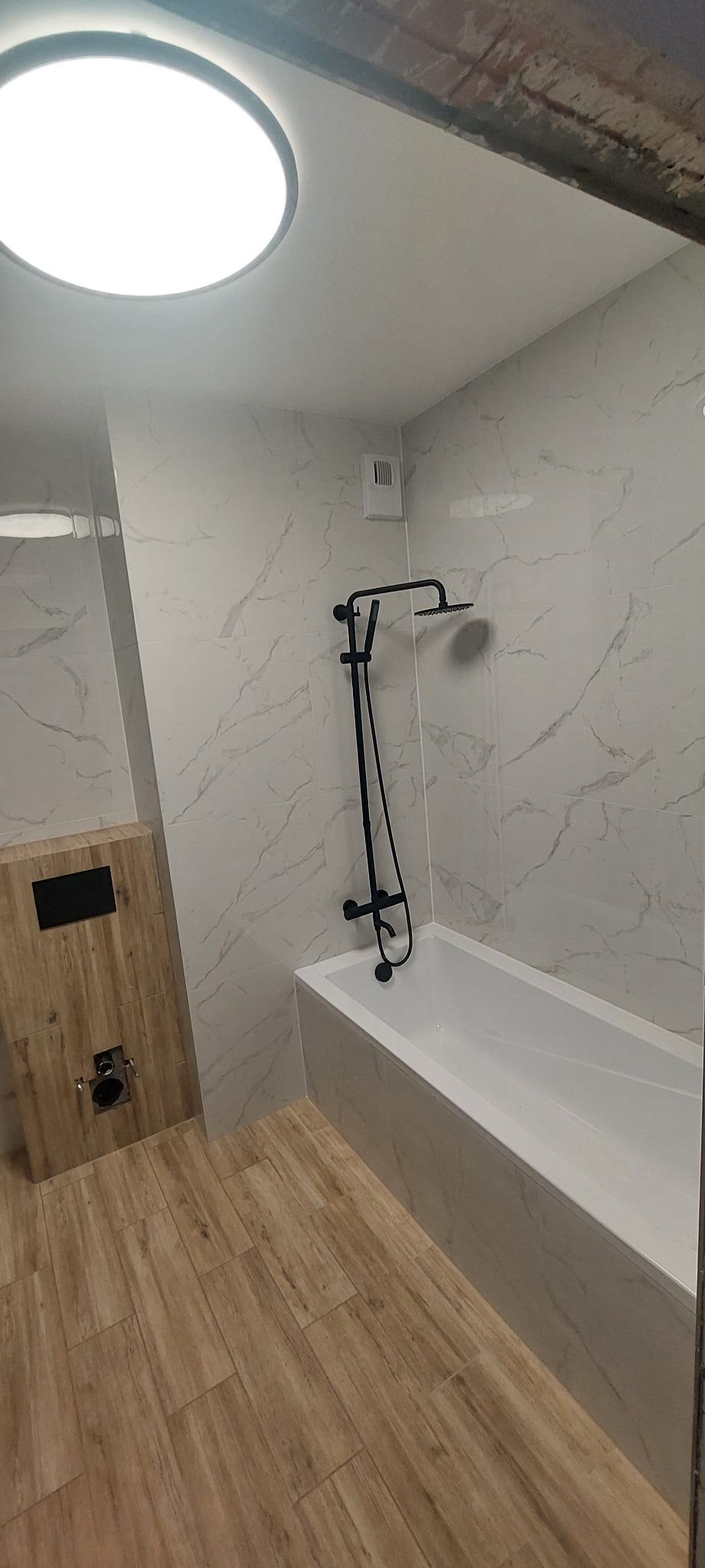 Wykończona łazienka z białą armaturą, czarną baterią prysznicową i płytkami imitującymi marmur na ścianach, drewnopodobna zabudowa WC.