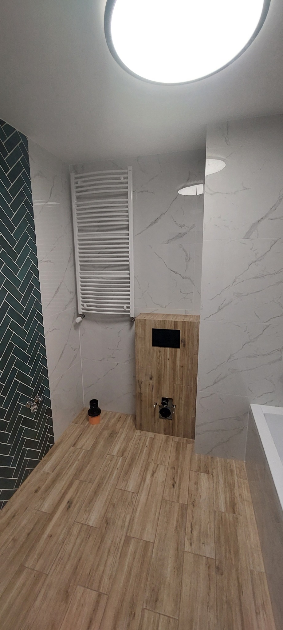Nowoczesna łazienka w trakcie wykończenia: zielona mozaika jodełka pod prysznicem, imitacja marmuru na ścianach, drewnopodobna podłoga, zabudowa WC z drewnianą okleiną i grzejnik drabinkowy.