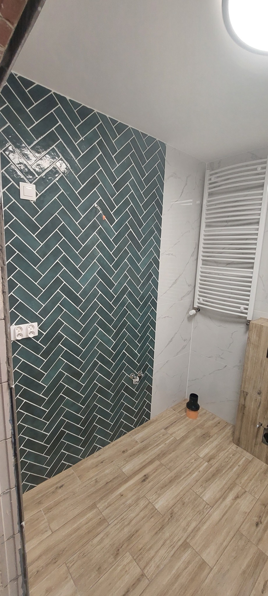 Ściana łazienki wyłożona ciemnozielonymi płytkami w jodełkę, z widocznymi gniazdkami elektrycznymi i fragmentem grzejnika drabinkowego na tle marmurowych płytek.