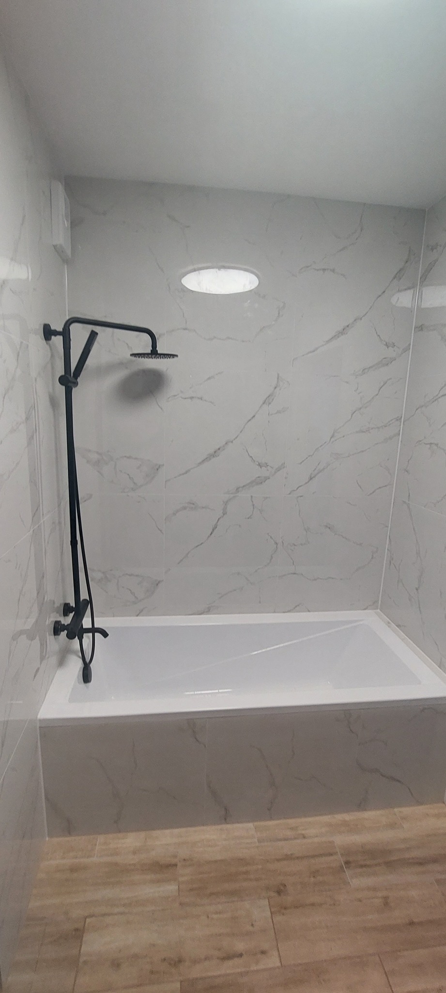 Nowoczesna łazienka po remoncie: biała wanna obudowana płytkami imitującymi marmur, czarna armatura natryskowa, oświetlenie sufitowe punktowe i podłoga wyłożona panelami imitującymi drewno.