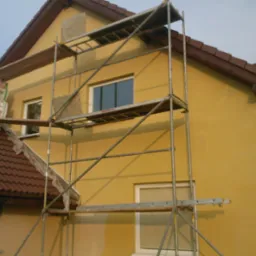 Żółta elewacja domu w trakcie prac, widoczne rusztowanie, styropian na dachu, fragment tynkowanej ściany i zakryte okno.
