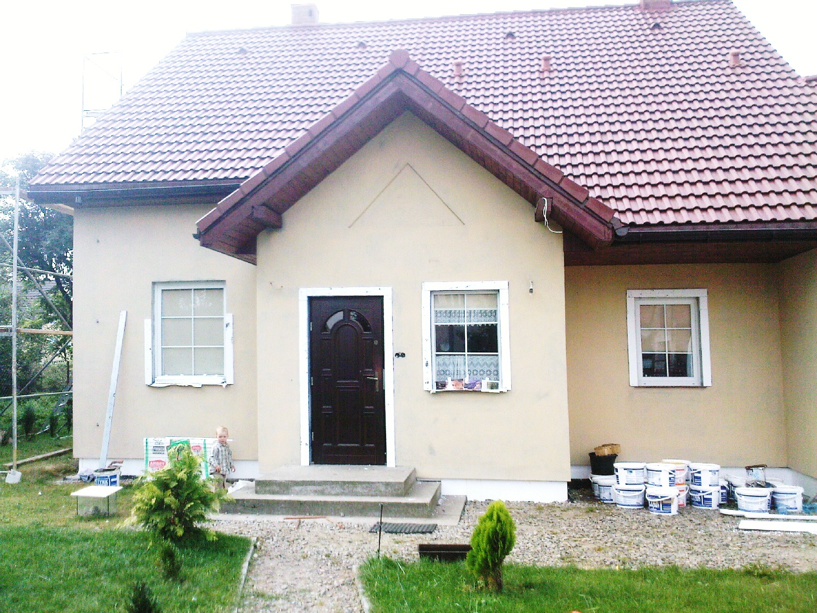 Dom jednorodzinny po nałożeniu tynku elewacyjnego, widoczne puszki z farbą, mały chłopiec i rusztowanie w tle.