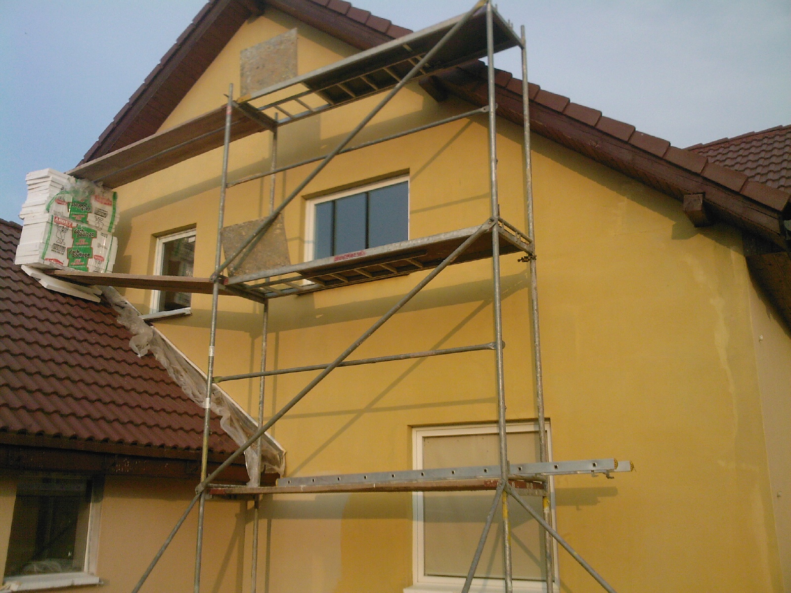 Żółta elewacja domu w trakcie prac, widoczne rusztowanie, styropian na dachu, fragment tynkowanej ściany i zakryte okno.