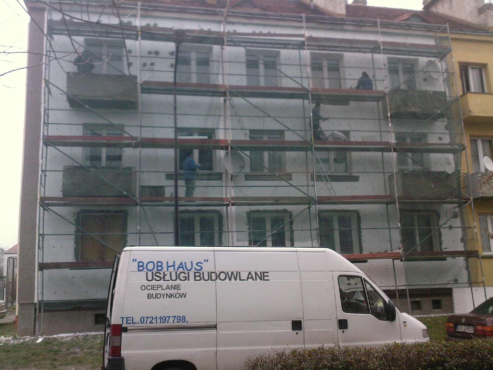 Wielopiętrowy budynek w trakcie ocieplania, widoczne rusztowania, częściowo ocieplona elewacja z szarymi panelami izolacyjnymi, na pierwszym planie biały samochód dostawczy z napisem 'BOB HAUS...