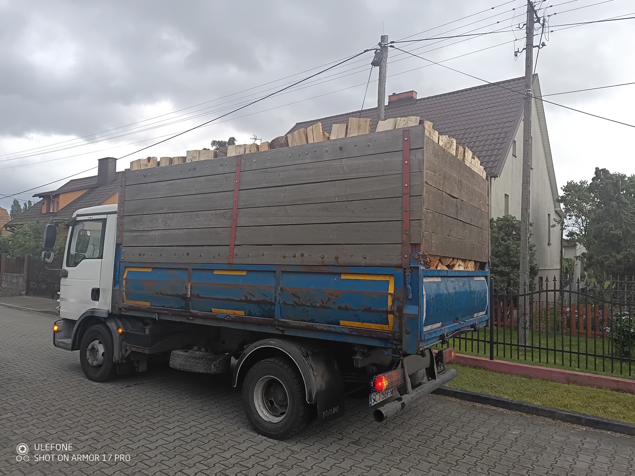 Ciężarówka MAN z pełną skrzynią drewna kominkowego na tle budynków mieszkalnych. Widok z boku, pochmurne niebo, kostka brukowa.