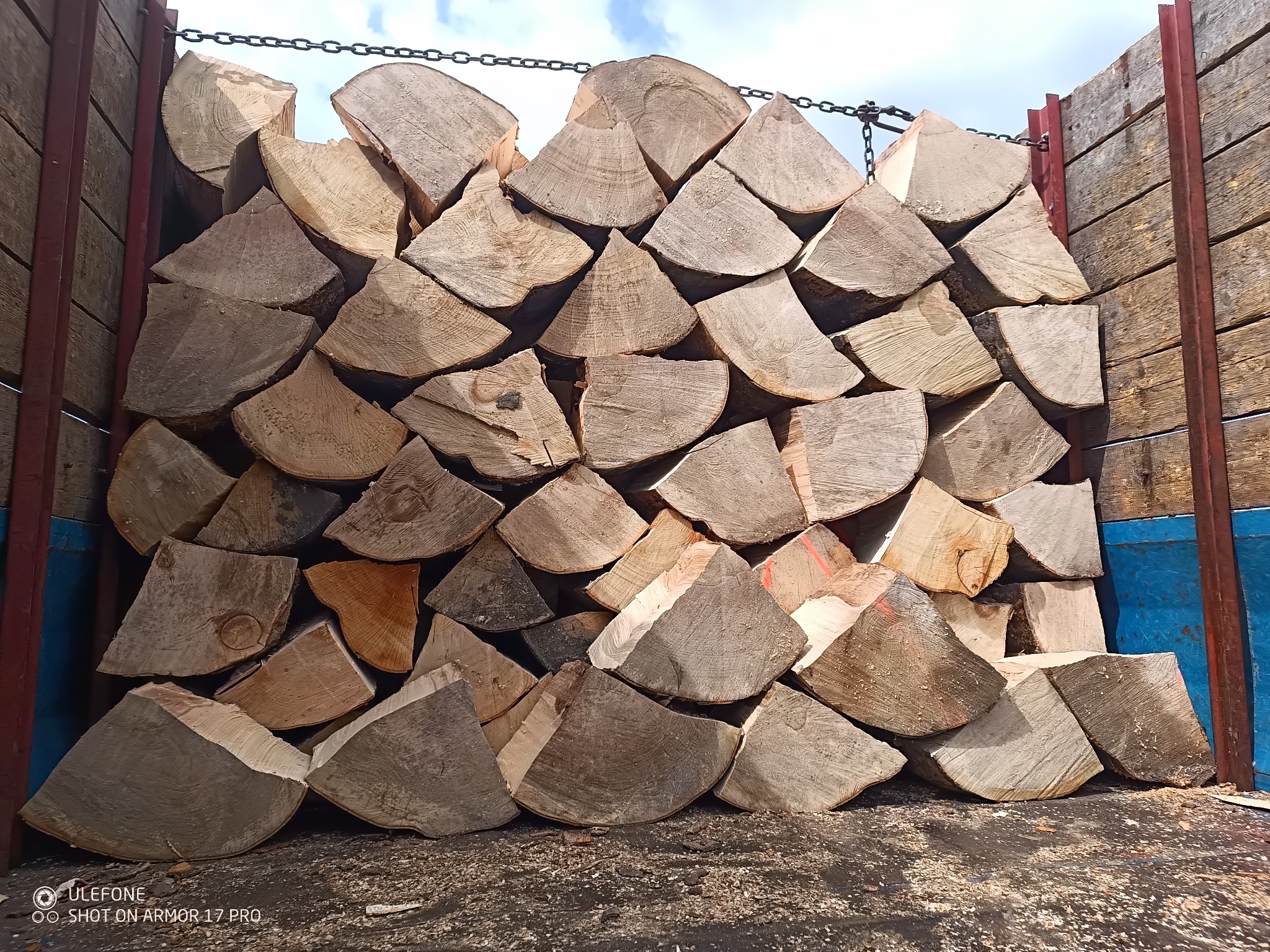 Ułożone w skrzyni kawałki drewna kominkowego, widoczne słoje i pęknięcia, zabezpieczone łańcuchem. Świeże drewno w transporcie, gotowe do sezonowania.