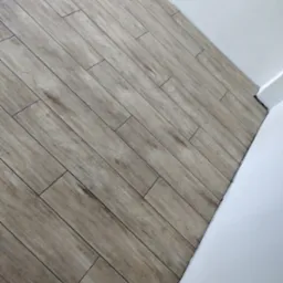 Ułożona pod kątem podłoga z płytek imitujących drewno, widoczna przy białej ścianie w rogu pomieszczenia.