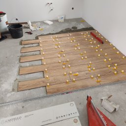 Usługi remontowo-budowlane - Układanie podłogi z płytek imitujących drewno, z użyciem poziomicy i klipsów poziomujących, widoczne paczki z płytkami Castello Ceramica i przecinarka do płytek Yato.