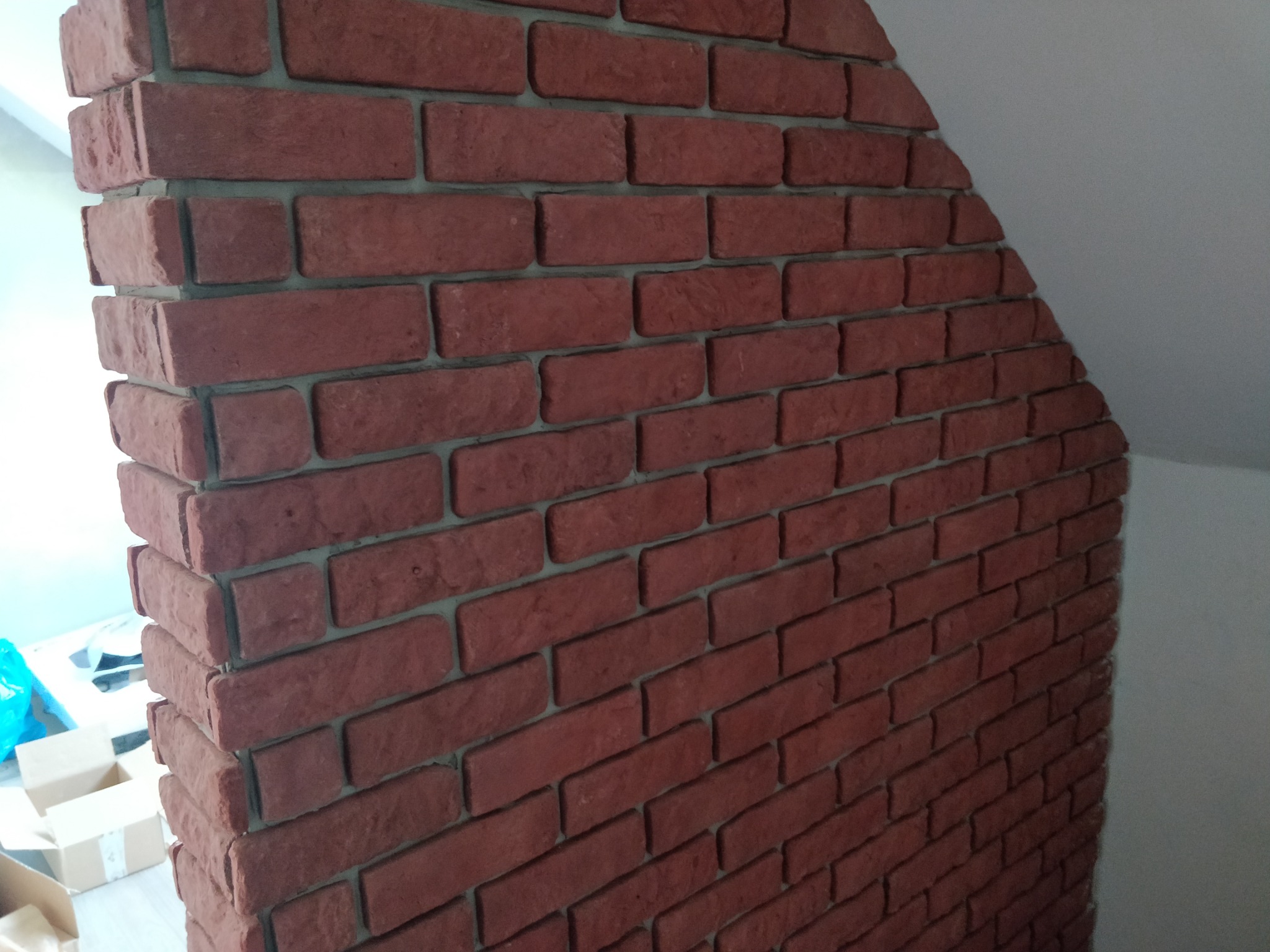 Ściana wykończona czerwoną cegłą klinkierową z szarą fugą, ułożona pod kątem na narożniku, widoczne kartony w tle.