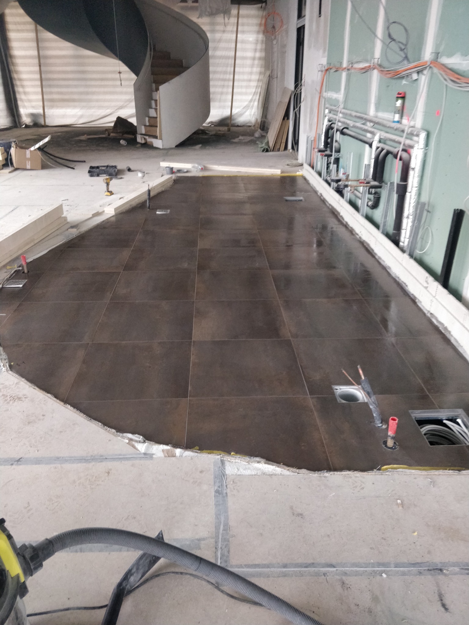 Układanie ciemnobrązowych płytek podłogowych w dużym pomieszczeniu w trakcie remontu, widoczne instalacje pod podłogą i schody spiralne w tle.