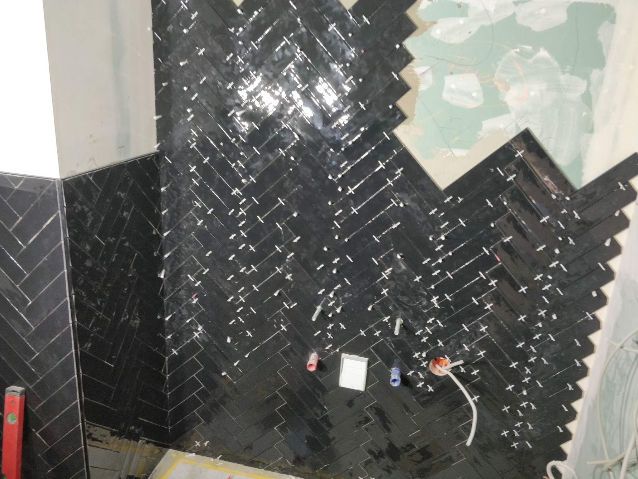Układanie czarnych, błyszczących płytek w jodełkę na ścianie, widoczne krzyżyki dystansowe, instalacja elektryczna i niezakończone fragmenty ściany z płyt kartonowo-gipsowych.