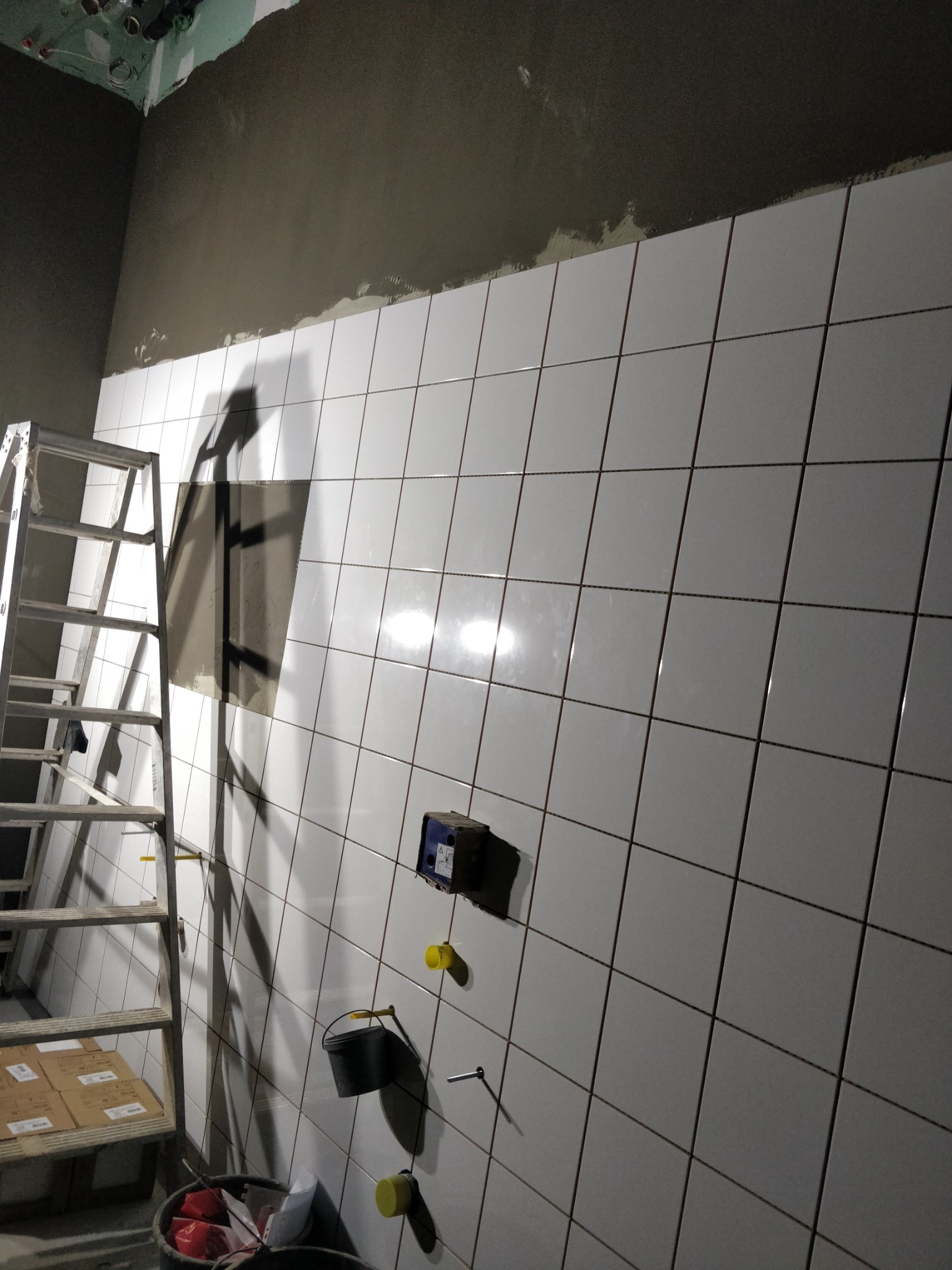 Ściana w trakcie kładzenia białych płytek, widoczny fragment surowego sufitu, drabina aluminiowa, puszka elektryczna i wiadro z narzędziami.