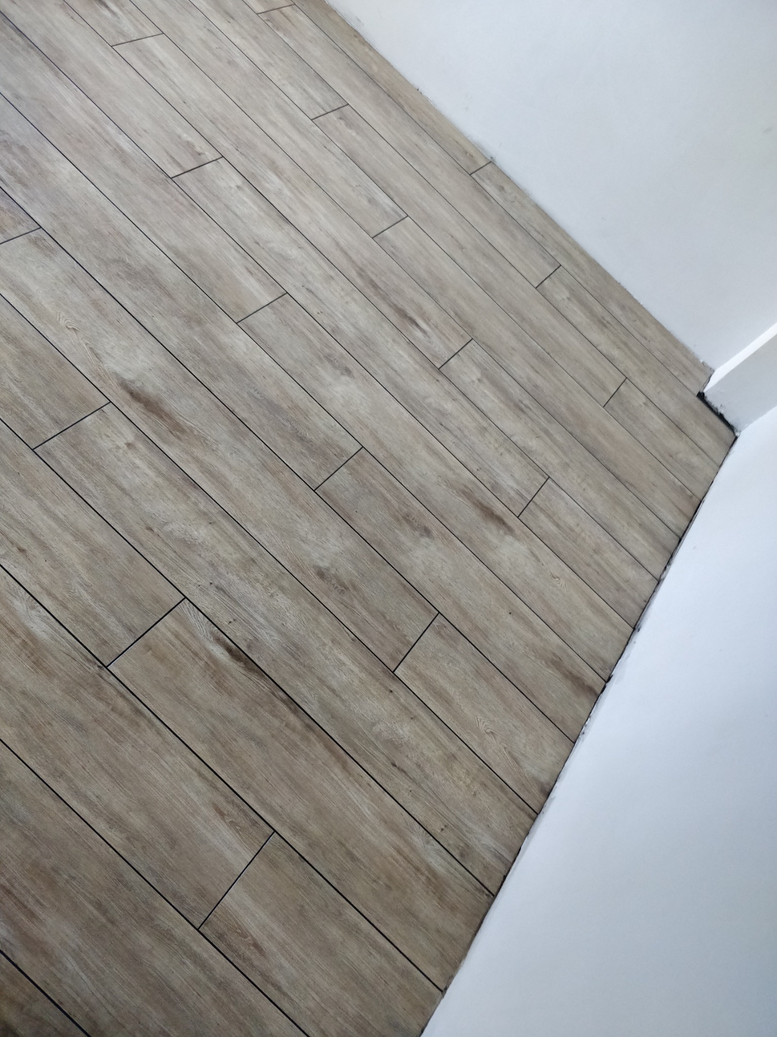 Ułożona pod kątem podłoga z płytek imitujących drewno, widoczna przy białej ścianie w rogu pomieszczenia.