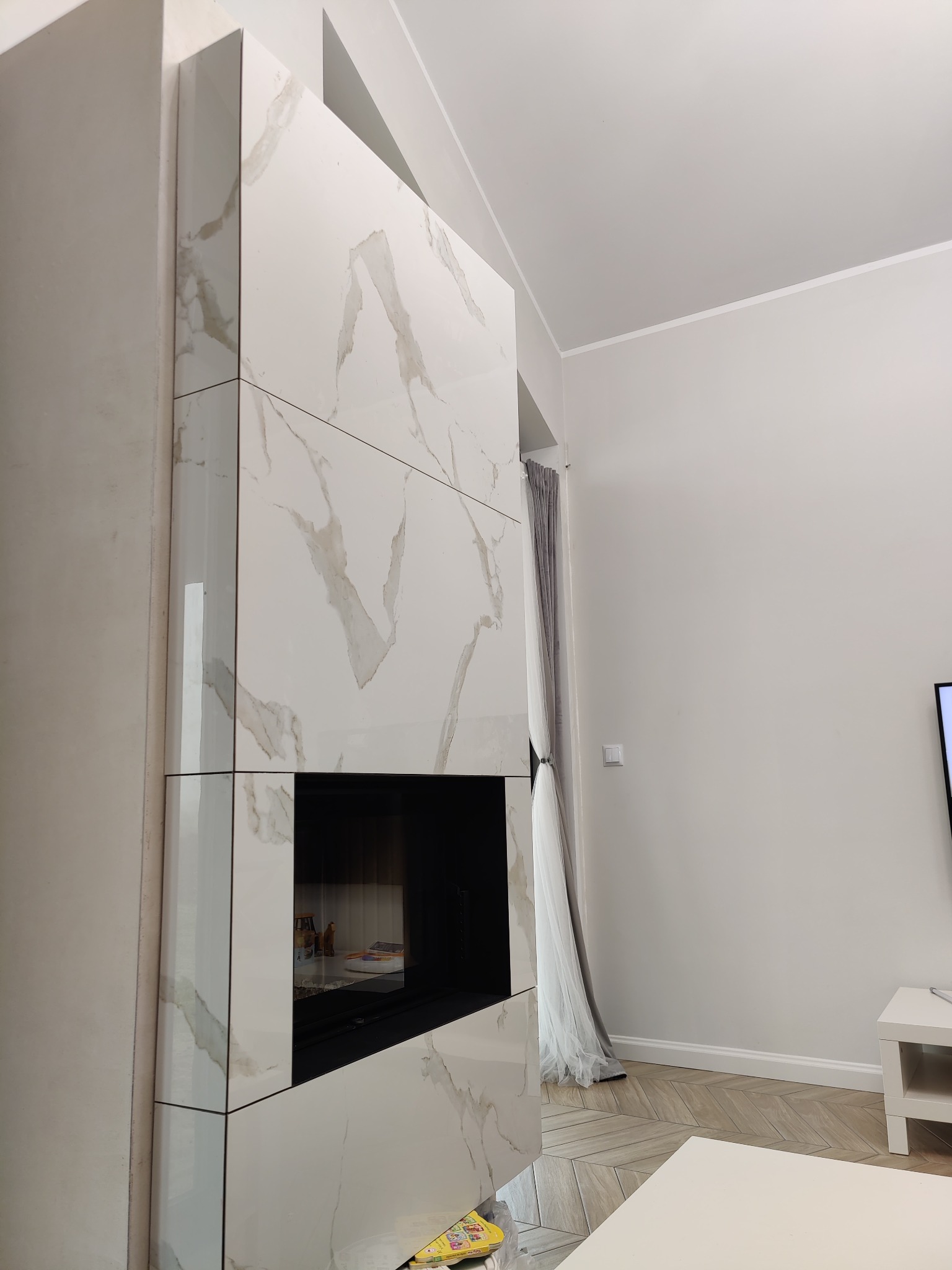 Wykończony kominek obudowany białymi, wielkoformatowymi płytami imitującymi marmur z widocznymi fugami, w nowoczesnym salonie z jasnymi ścianami i drewnianą podłogą ułożoną w jodełkę.