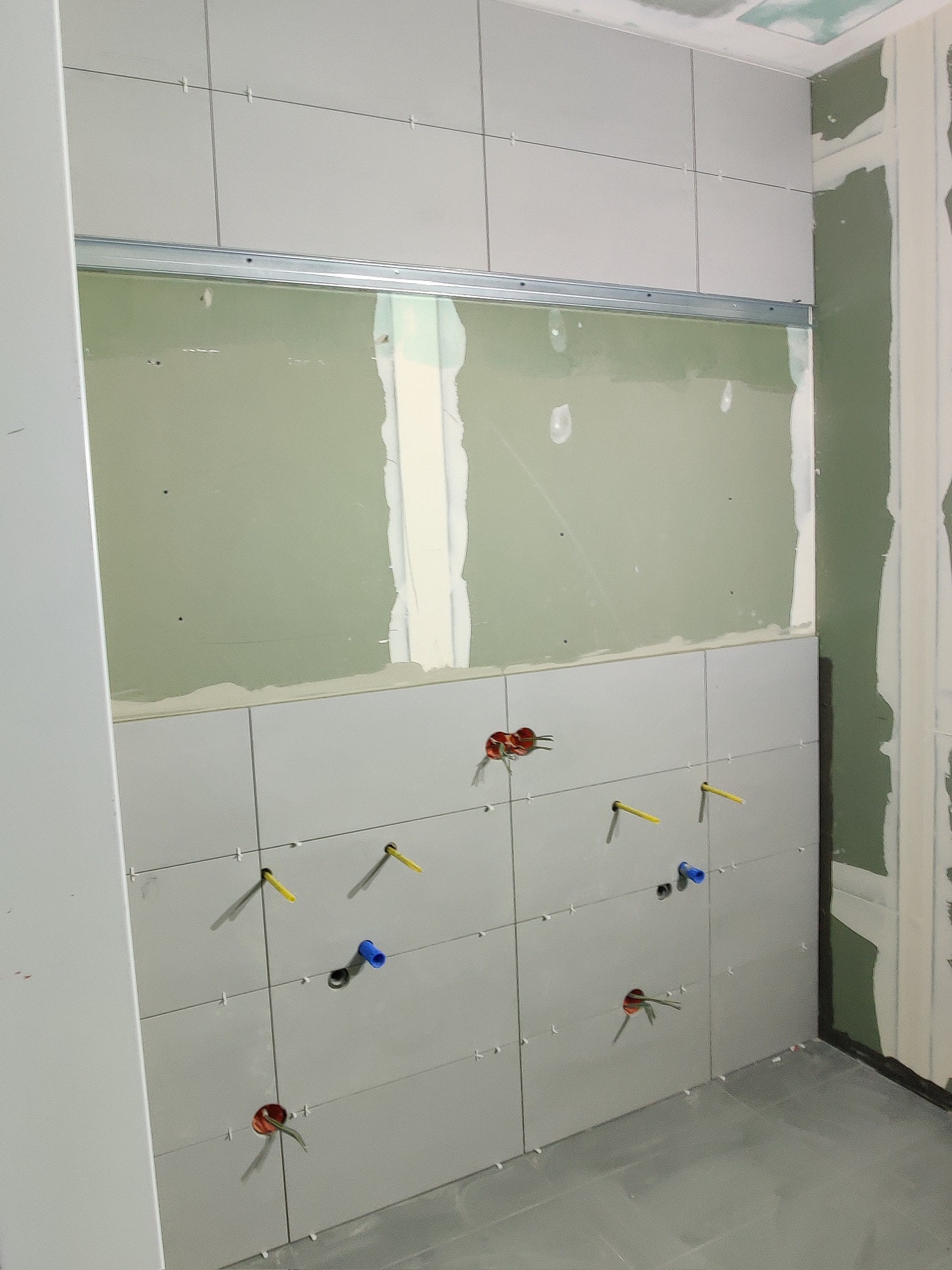Ściana łazienki w trakcie remontu: częściowo ułożone szare płytki, widoczne otwory na instalacje, powyżej płytek płyta kartonowo-gipsowa na metalowym profilu, ściana obok pokryta płytą...