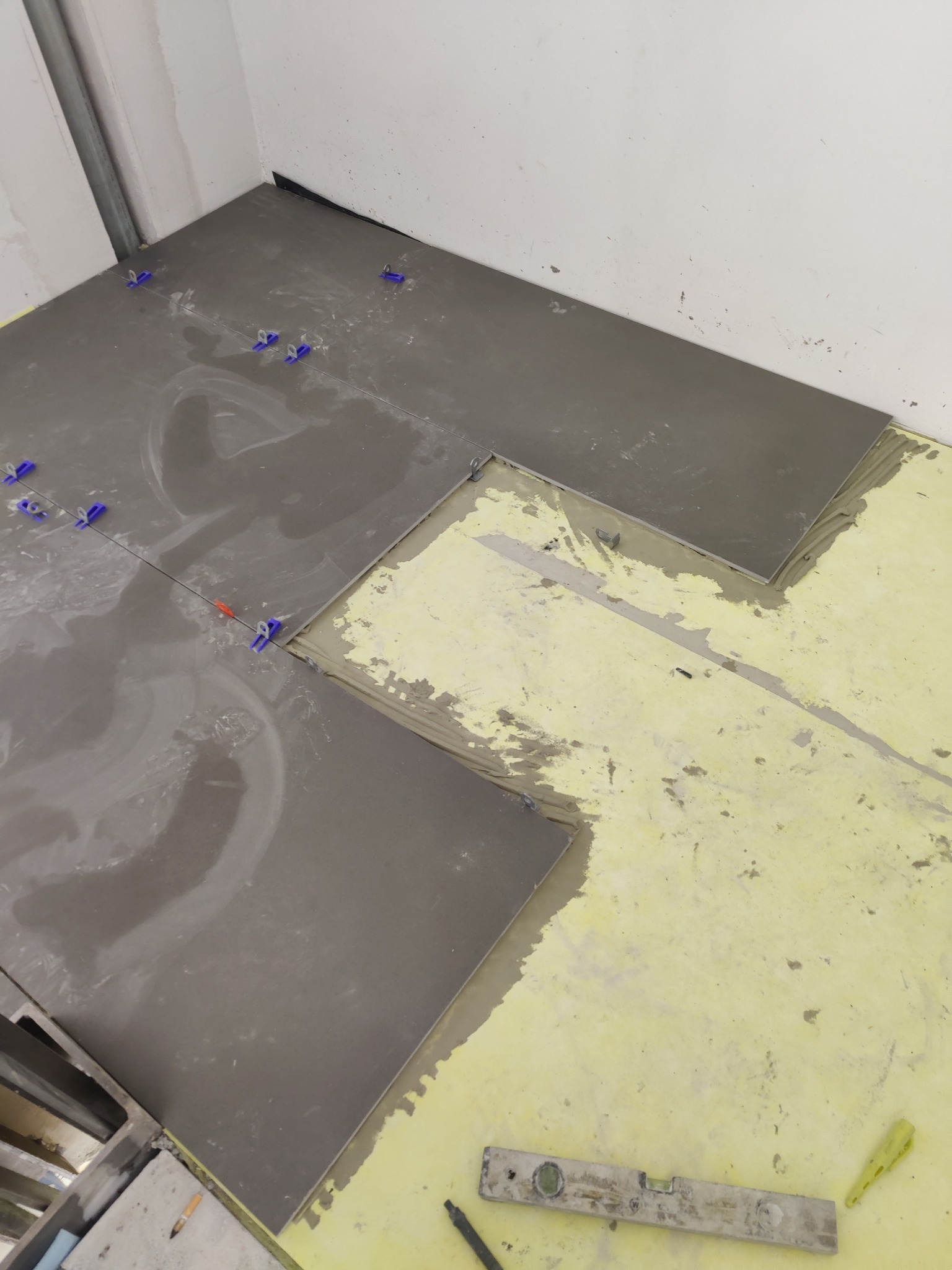 Układanie ciemnoszarych płytek podłogowych z użyciem niebieskich krzyżyków dystansowych, widoczna warstwa kleju na żółtej macie podkładowej oraz poziomica i ołówek na podłodze.