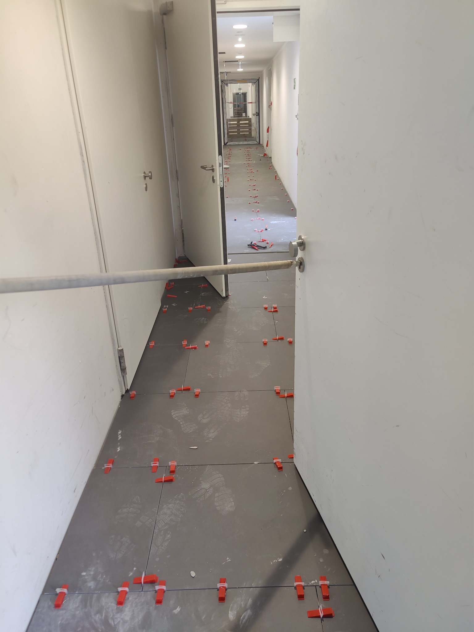 Korytarz w trakcie układania szarych płytek, widoczne ślady butów i czerwone klipsy poziomujące, drzwi po obu stronach.