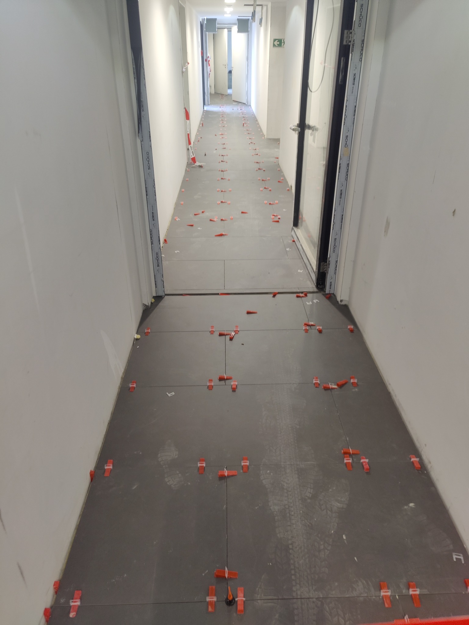 Korytarz w trakcie układania ciemnoszarych płytek podłogowych z użyciem czerwonych klipsów poziomujących; widoczne ślady butów na powierzchni płytek.