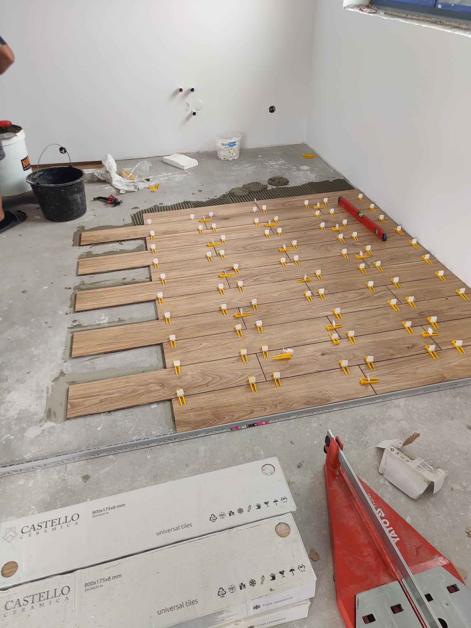 Układanie podłogi z płytek imitujących drewno, z użyciem poziomicy i klipsów poziomujących, widoczne paczki z płytkami Castello Ceramica i przecinarka do płytek Yato.