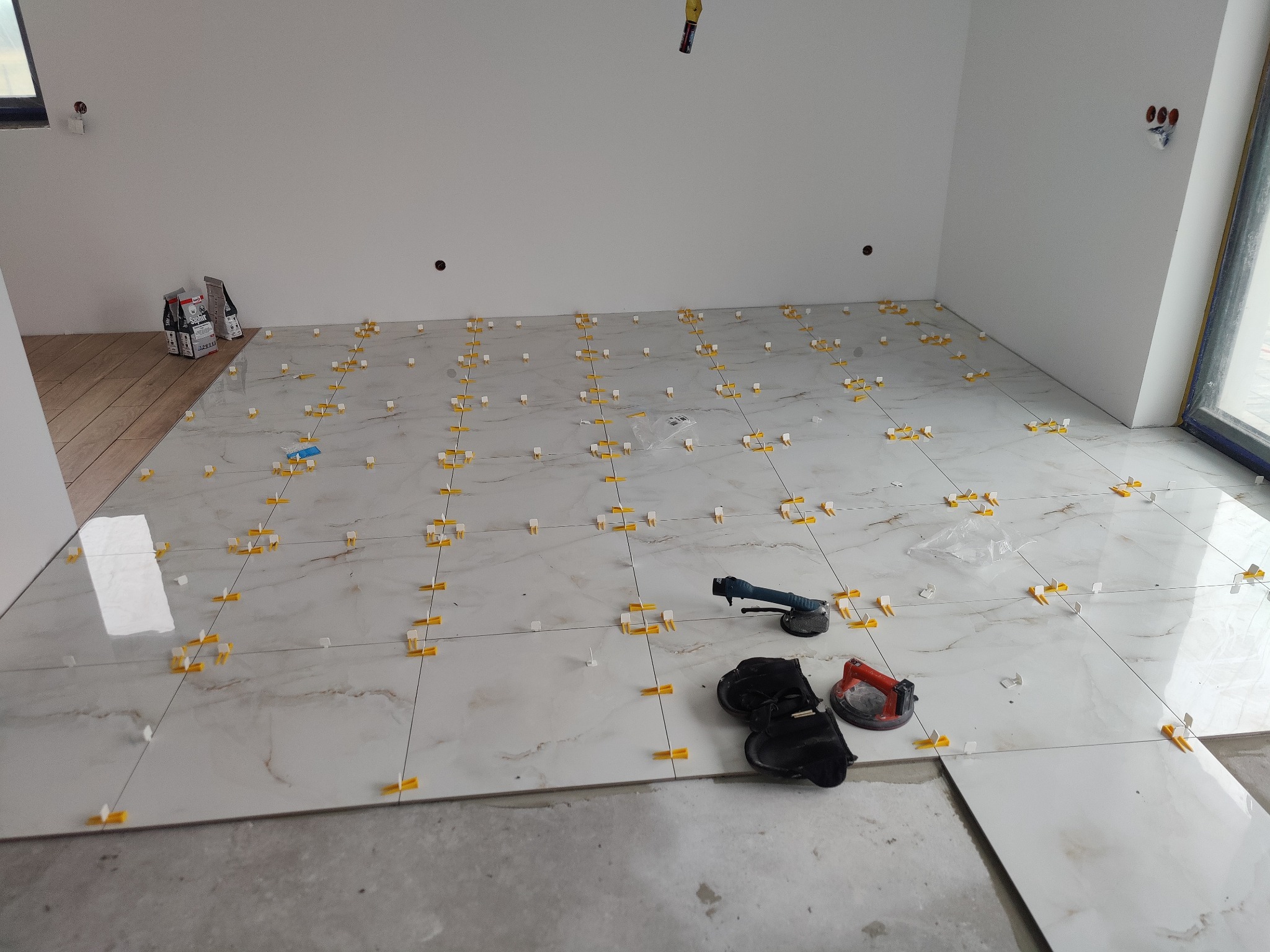 Układanie płytek podłogowych w jasnym pomieszczeniu, widoczne narzędzia glazurnicze i system poziomowania płytek z żółtymi klipsami.