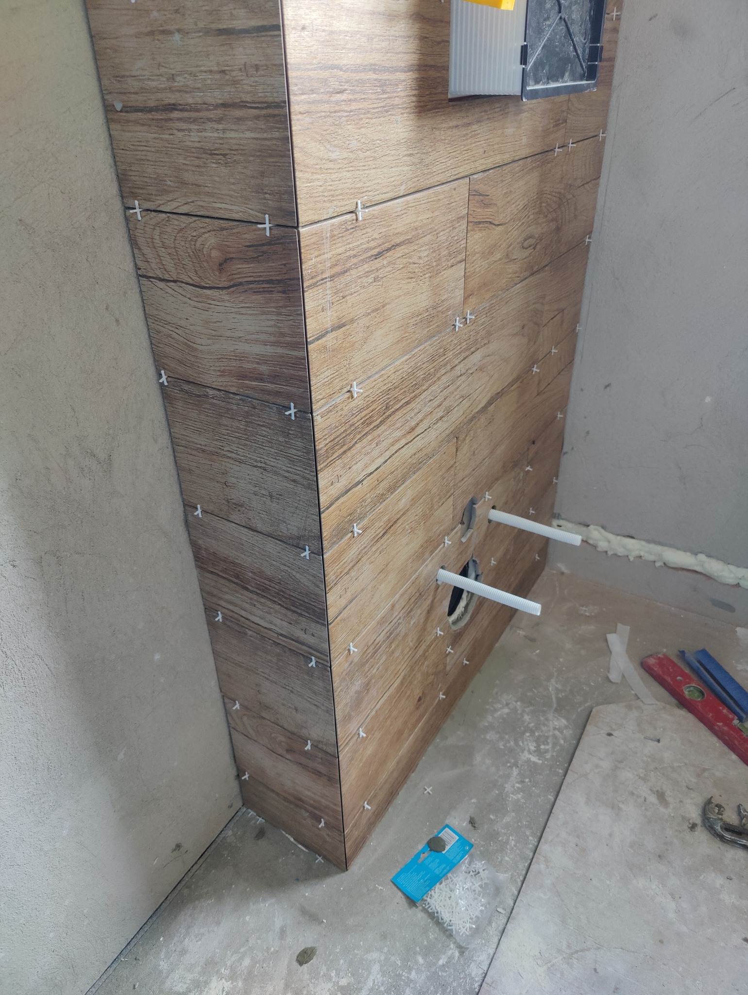 Obudowa stelaża podtynkowego WC wykończona płytkami imitującymi drewno, z widocznymi rurami i krzyżykami dystansowymi, w trakcie prac wykończeniowych.