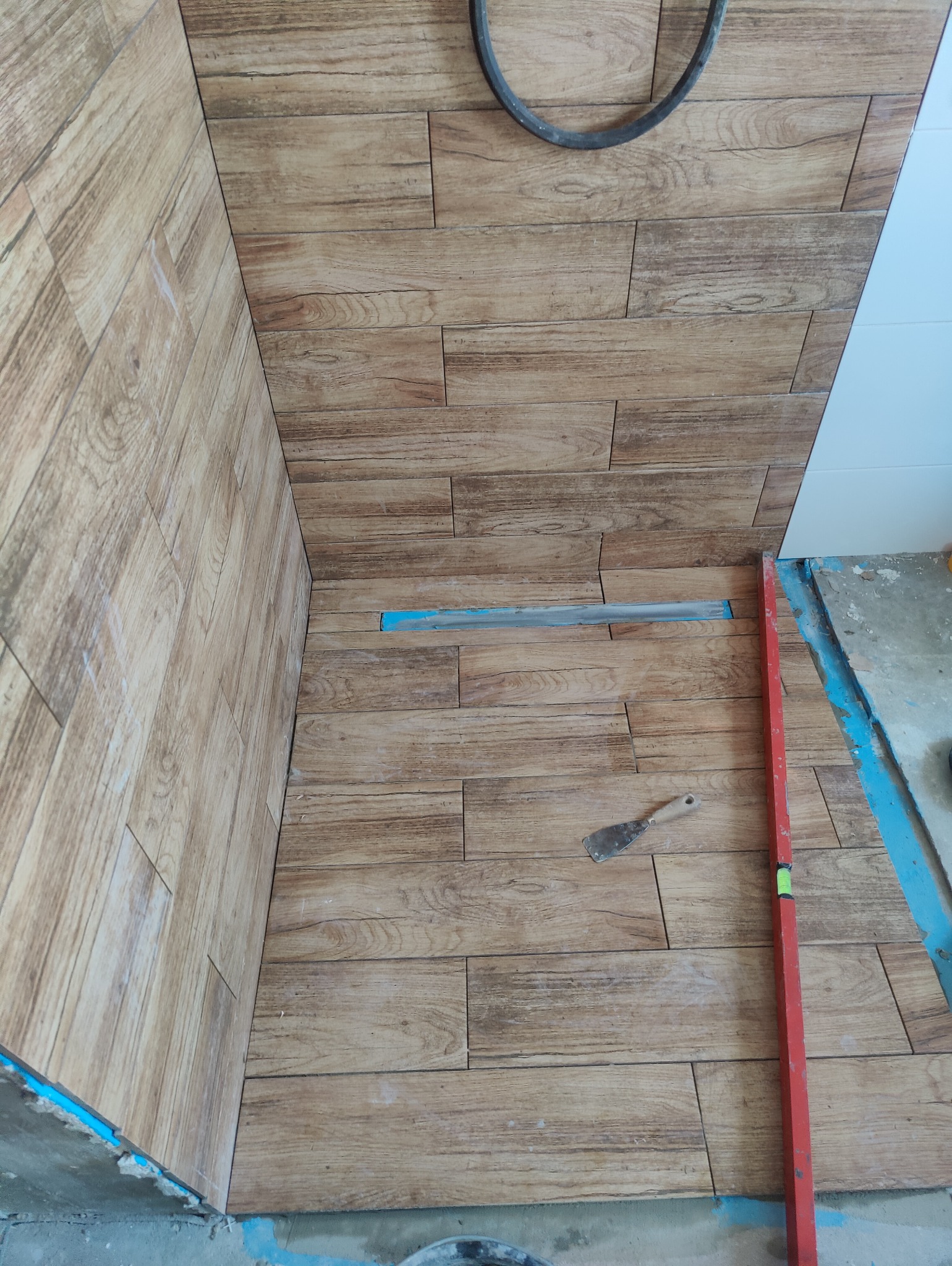 Układanie płytek imitujących drewno na podłodze i ścianie w łazience, widoczna poziomica i kielnia, nieukończony etap prac.