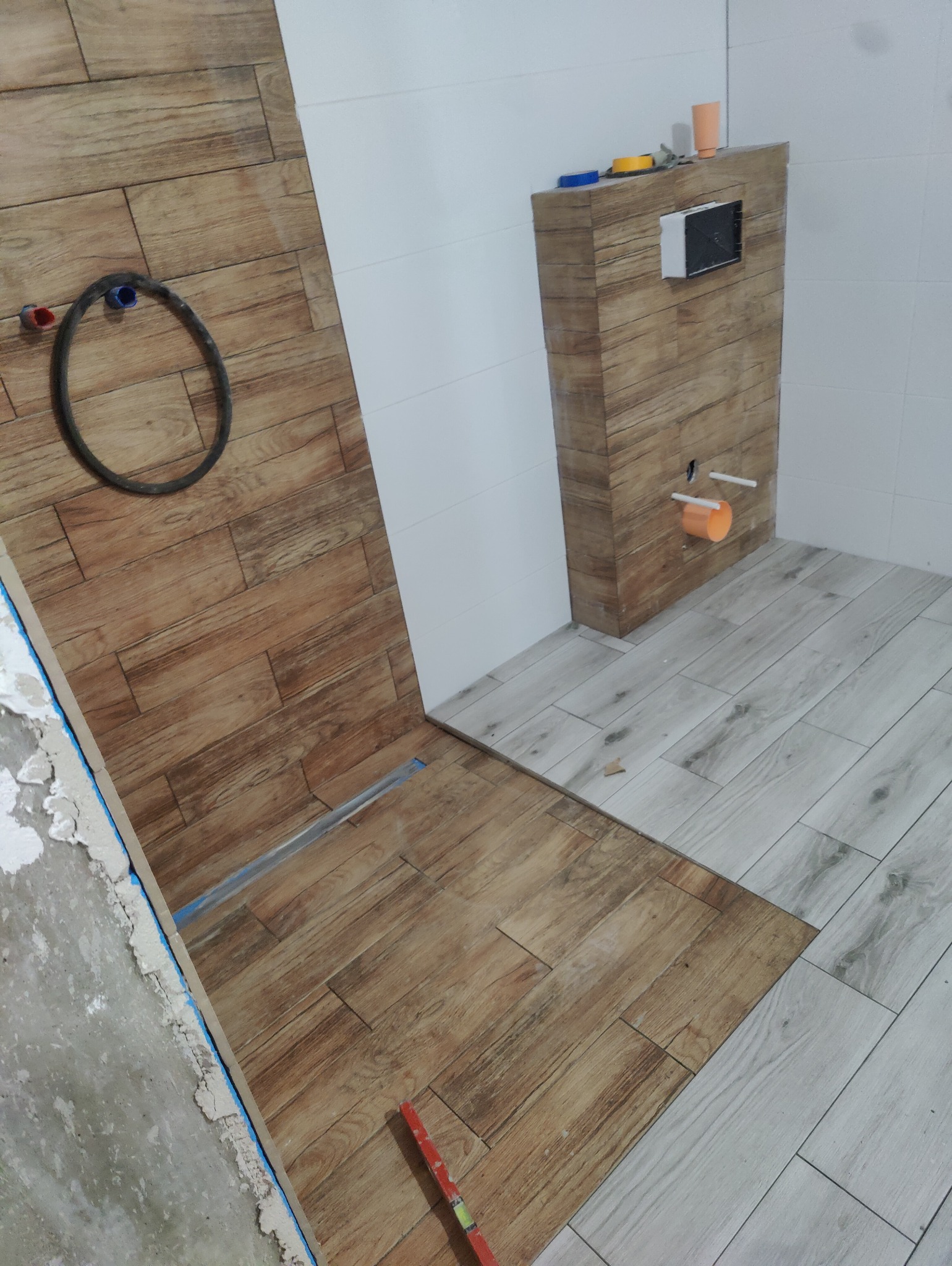 Łazienka w trakcie remontu, widoczne ułożone płytki imitujące drewno na ścianie i podłodze, instalacja podtynkowego WC oraz fragment ściany z surowym tynkiem.