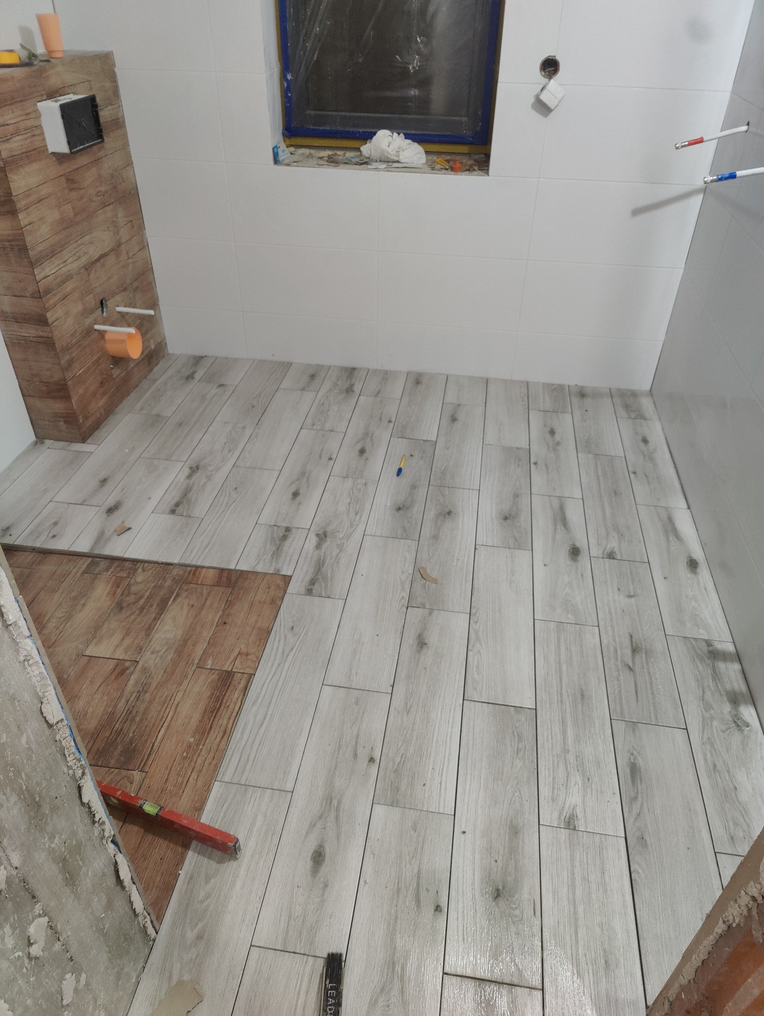 Układanie jasnych, drewnopodobnych płytek podłogowych w łazience. Częściowo ułożone płytki w odcieniach brązu. Widoczna poziomica i fragment ściany w trakcie prac wykończeniowych.