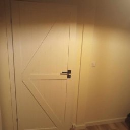 Daniel Więcek - Białe drzwi wewnętrzne w stylu rustykalnym z czarną klamką, zamontowane w jasnym pomieszczeniu z panelami podłogowymi.