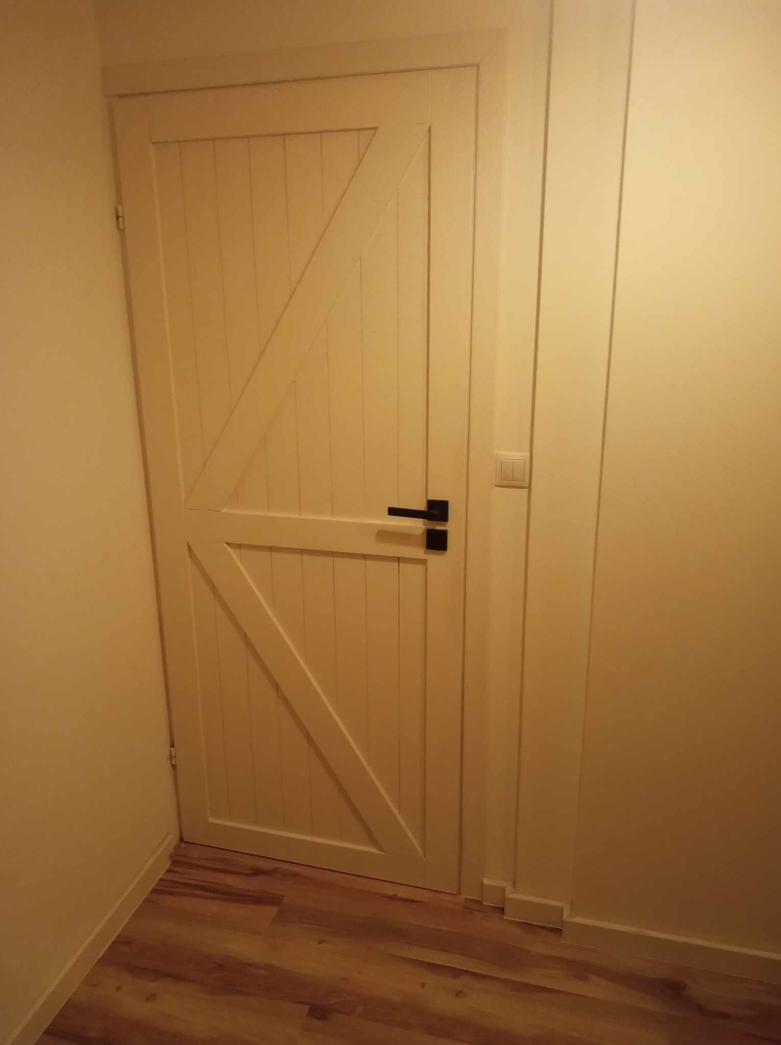 Zamknięte, kremowe drzwi wewnętrzne z ukośnymi elementami dekoracyjnymi i czarną klamką na tle jasnej ściany i podłogi imitującej drewno.