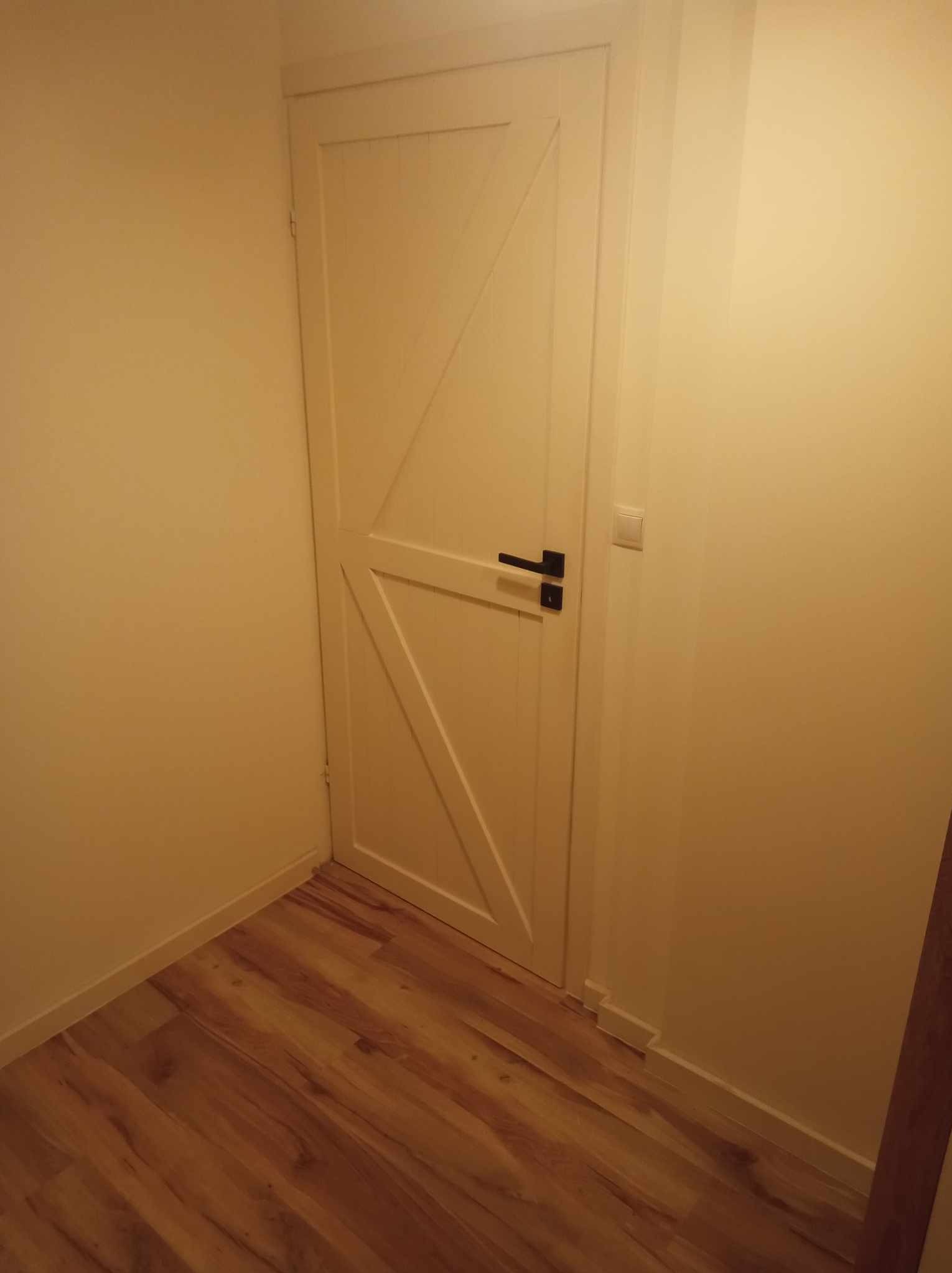 Wewnętrzna, biała, drewniana drzwi z czarną klamką, w korytarzu z panelami podłogowymi imitującymi drewno i jasnymi ścianami.