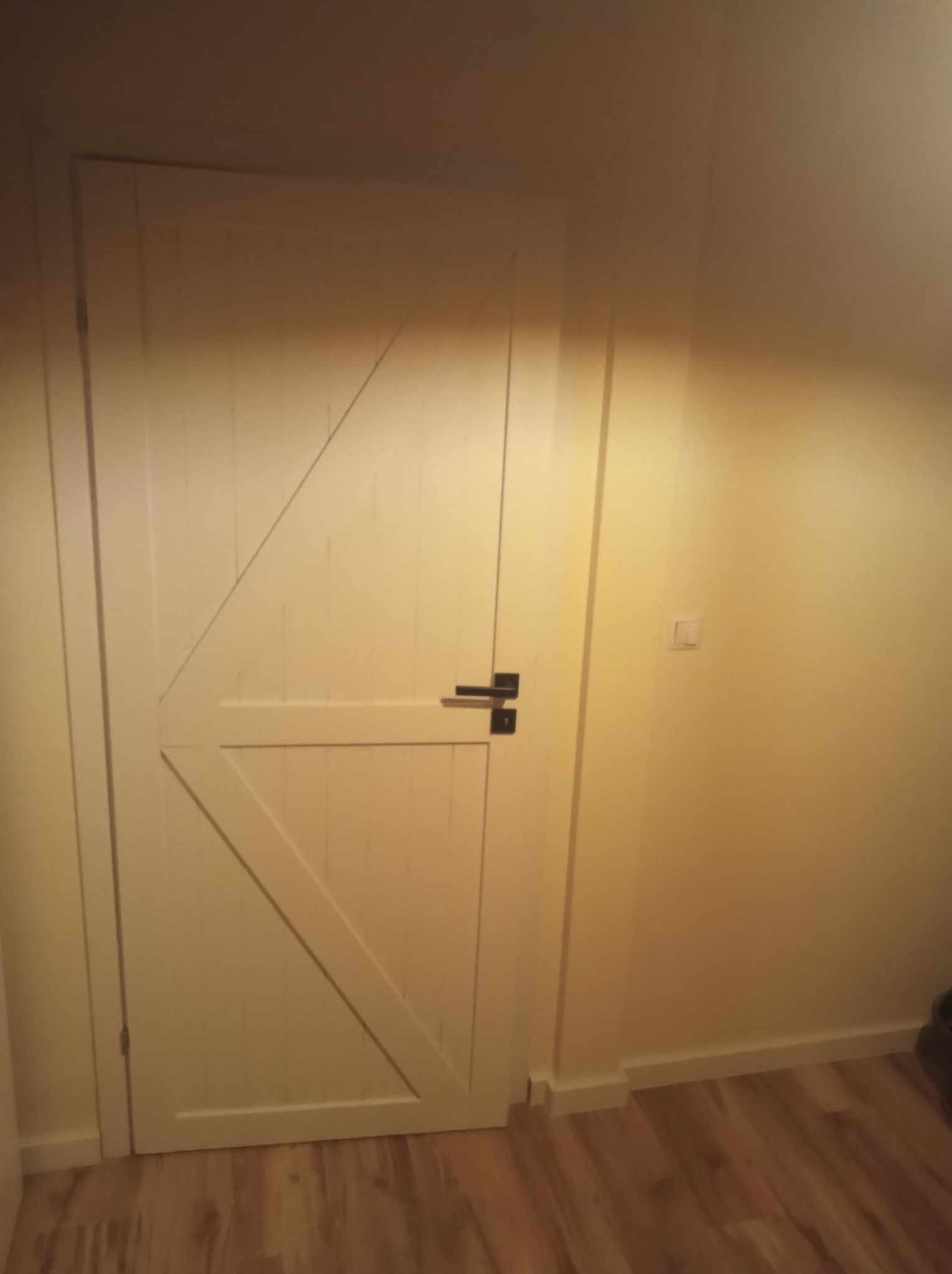 Białe drzwi wewnętrzne w stylu rustykalnym z czarną klamką, zamontowane w jasnym pomieszczeniu z panelami podłogowymi.